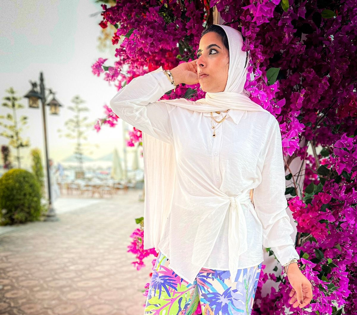 Woman in a white outfit with colorful pants standing among pink flowers.