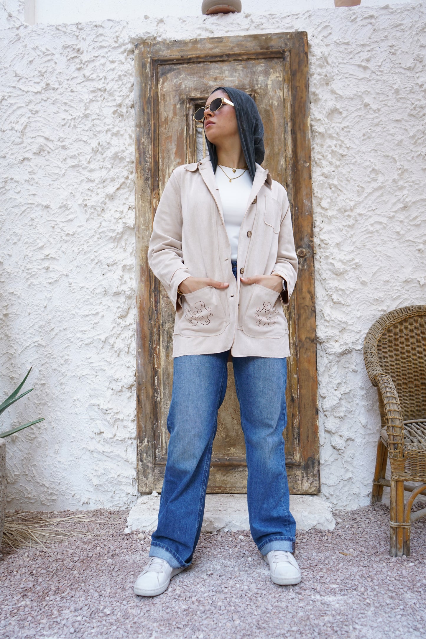 Person wearing a beige coat and blue jeans standing in front of a textured wall with a wooden door.
