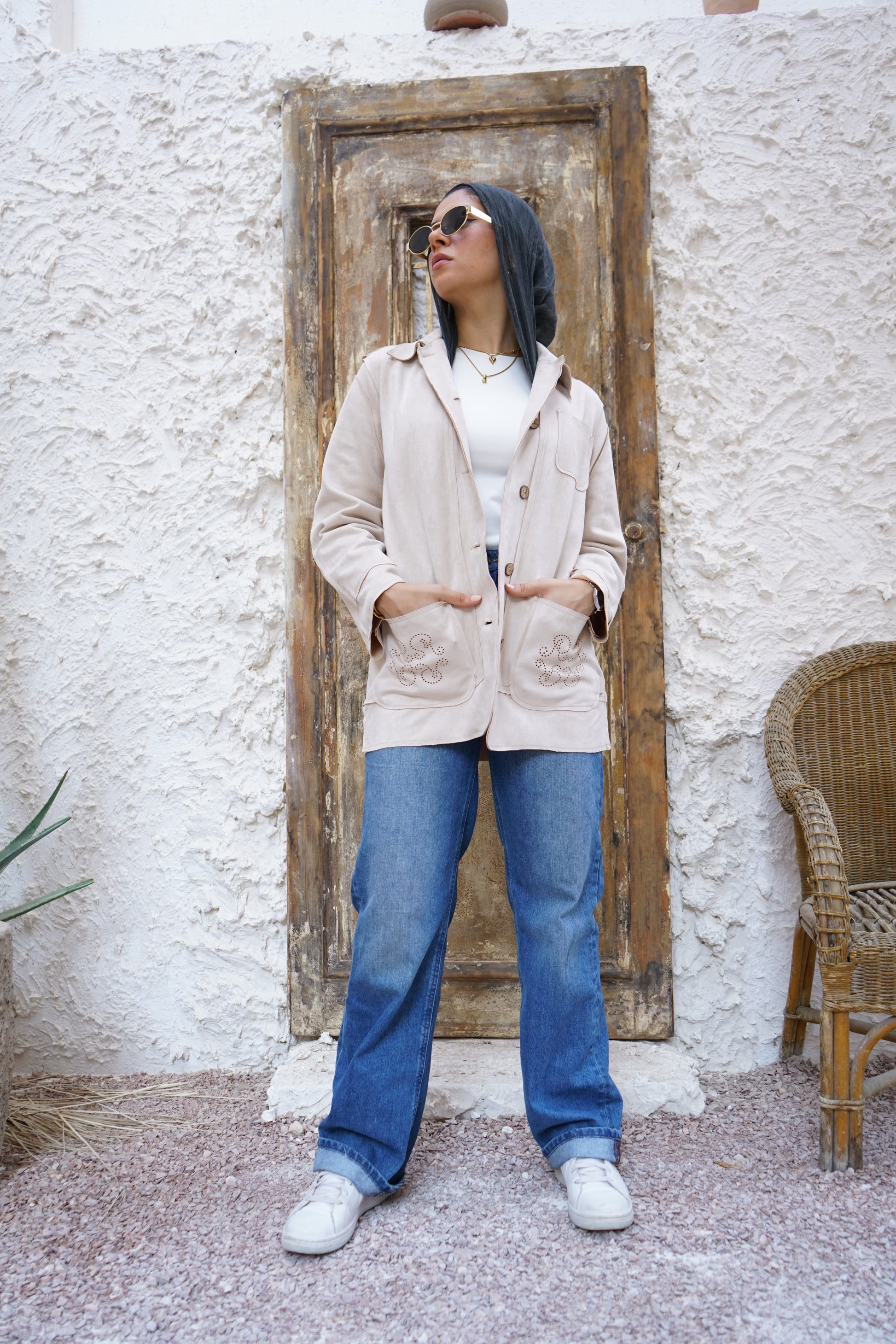 Person wearing a beige coat and blue jeans standing in front of a textured wall with a wooden door.